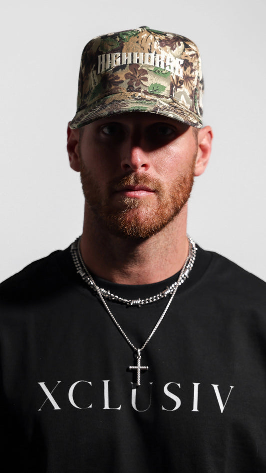 Man wearing a camouflage cap and black 'XCLUSIV' shirt on a gray background